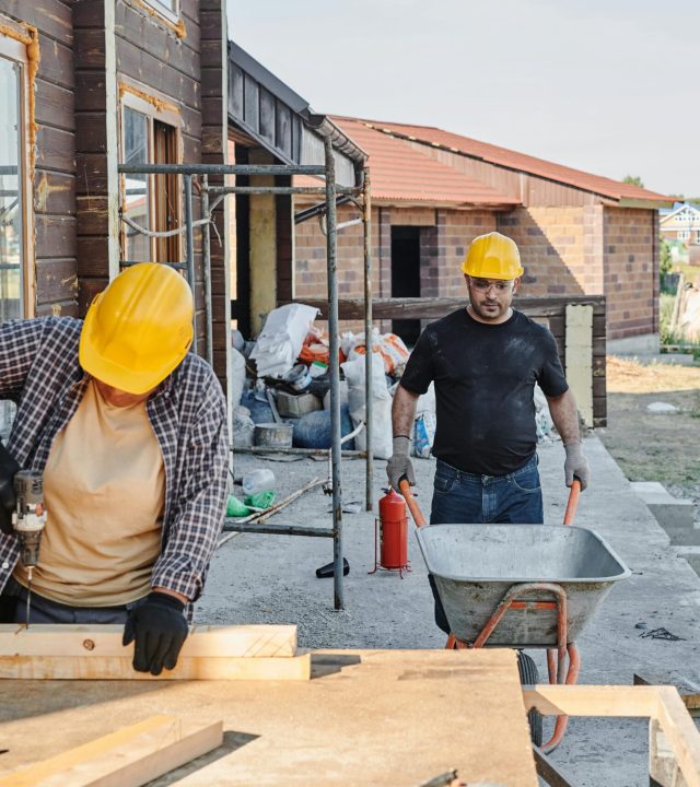 Construction workers building a house, using a saw and carrying materials in a wheelbarrow.